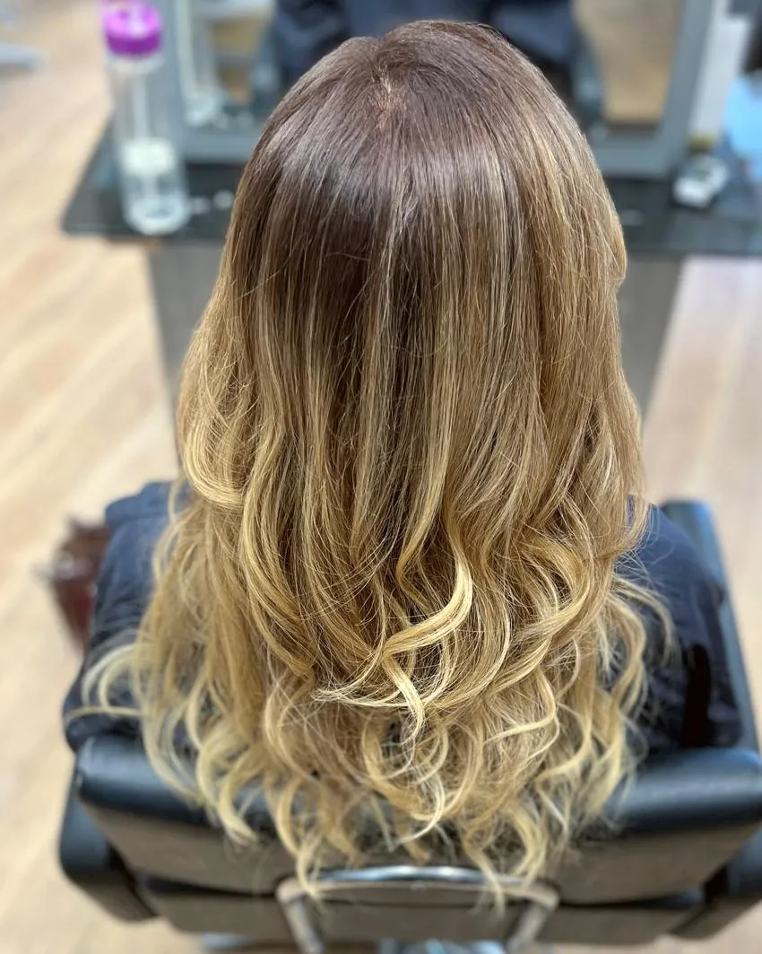 Long hair running from a rich brown root into warm golden-blonde ends, set in loose, bouncy curls.